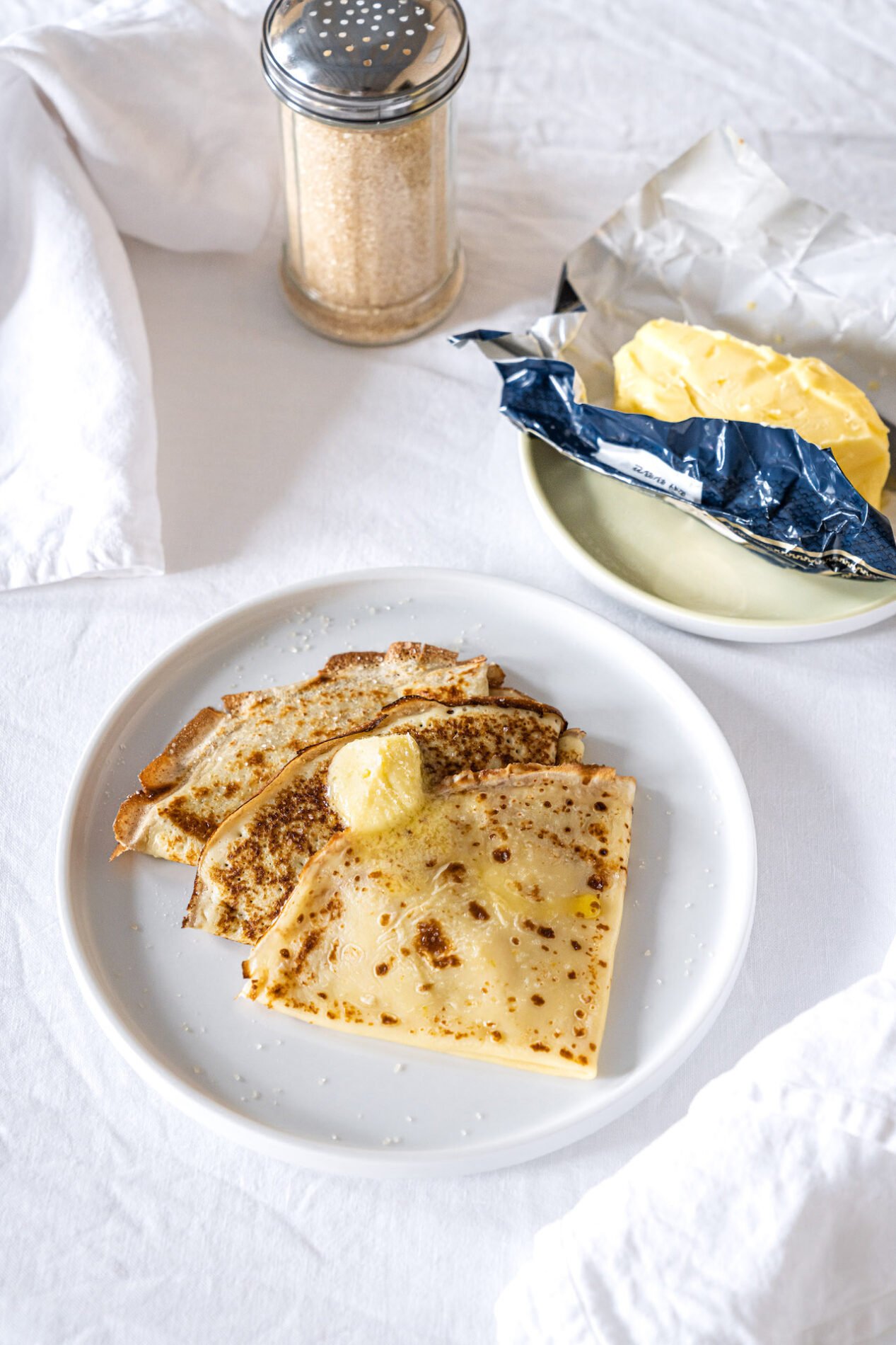 Trois crêpes dans une assiette avec une noisette de beurre, avec une plaquette de beurre et un pot sucre